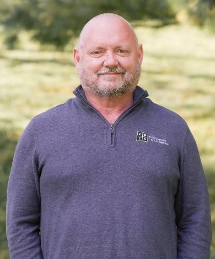 Smiling bald man in a purple quarter-zip sweater standing outdoors with a blurred grassy background