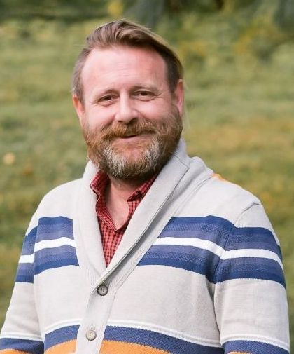 Smiling man in a beige cardigan with blue stripes and a red checkered shirt, outdoors in front of blurred greenery