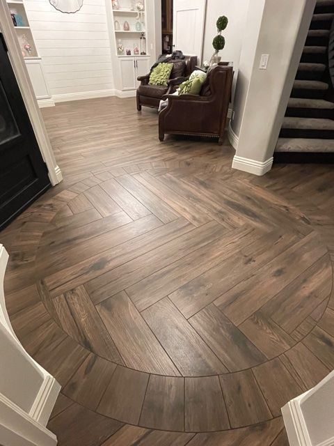 Brown wooden herringbone-patterned floor in a home's entryway, with a curved border and chairs in the background.