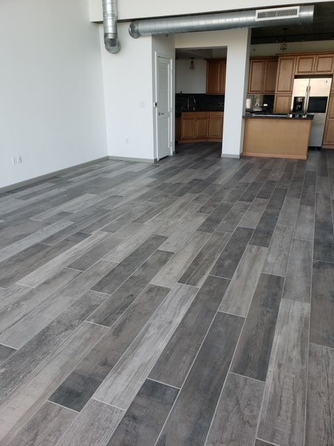 Gray wood-look tile flooring in a modern apartment, leading towards a kitchen with brown cabinets.