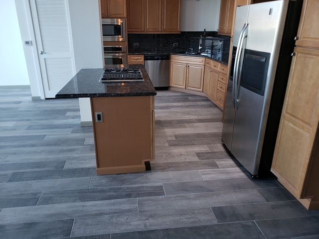 Kitchen with wood cabinets, stainless steel appliances, island with stovetop, and gray wood-look flooring.
