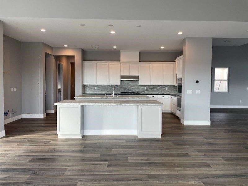 Spacious kitchen with white cabinets, gray backsplash, and island. Dark wood floor, and gray walls.