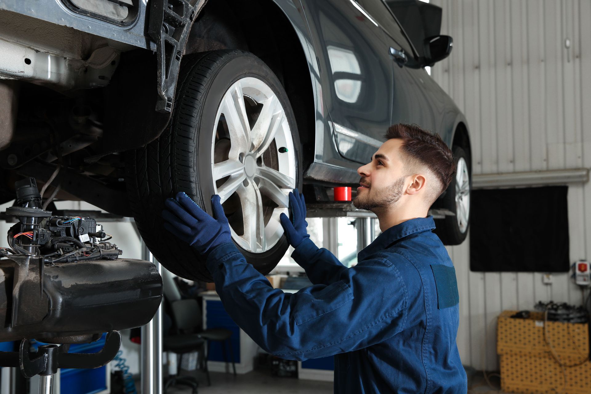 A technician in blue coveralls and gloves inspecting a car tire raised on a lift in a repair shop.