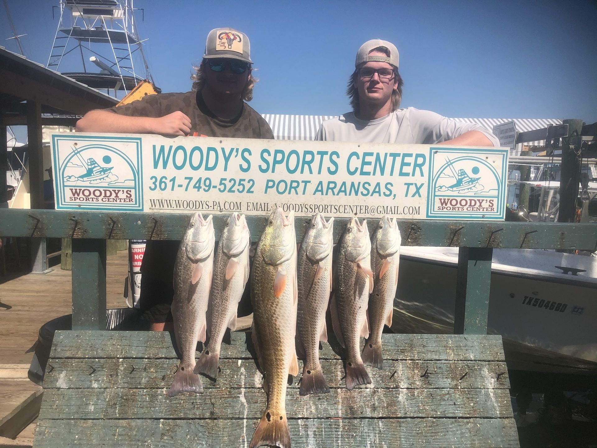 Two men are holding a bunch of fish in front of a woody 's sports center sign.