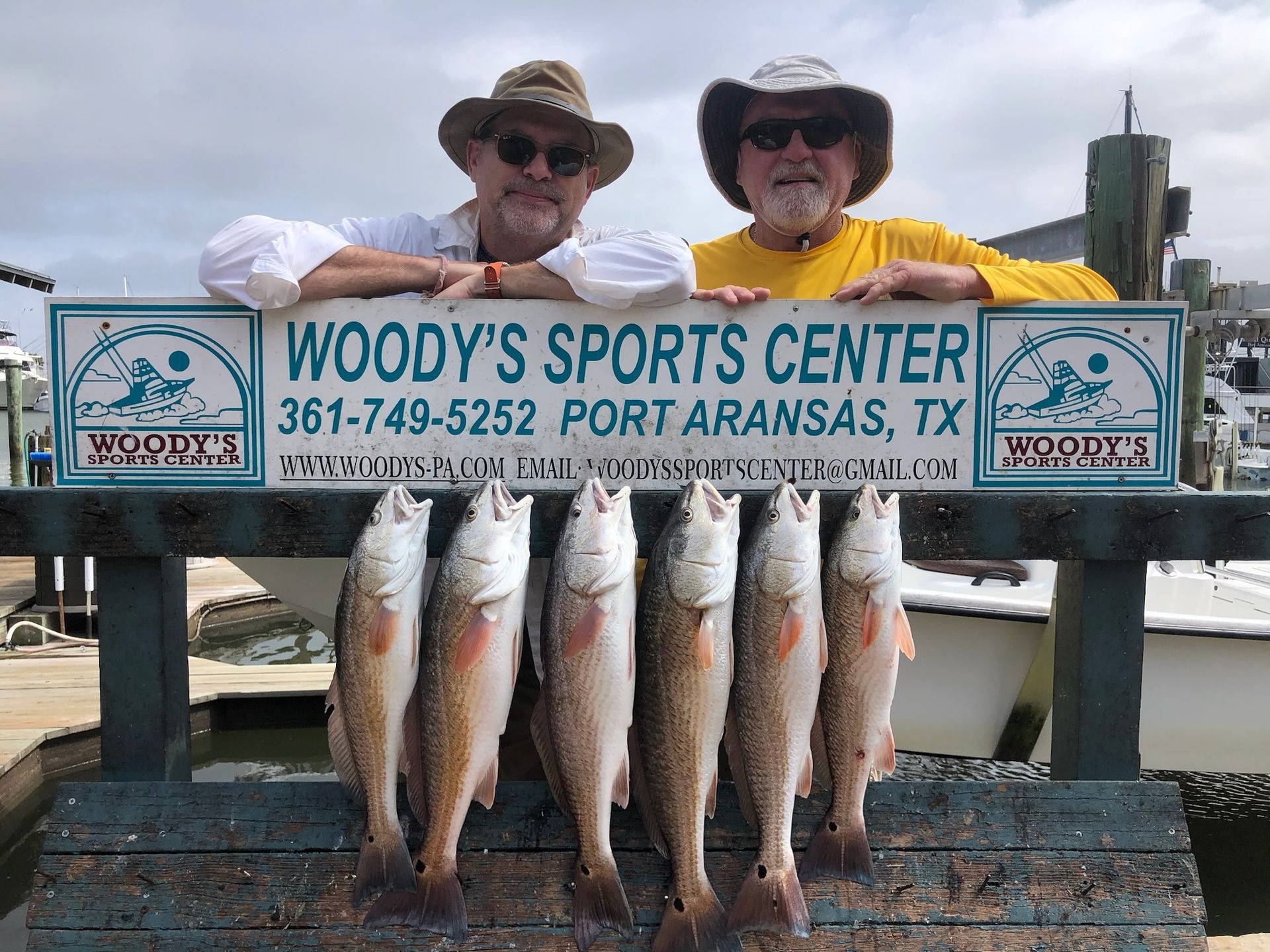 Two men are holding a bunch of fish in front of a woody 's sports center sign.