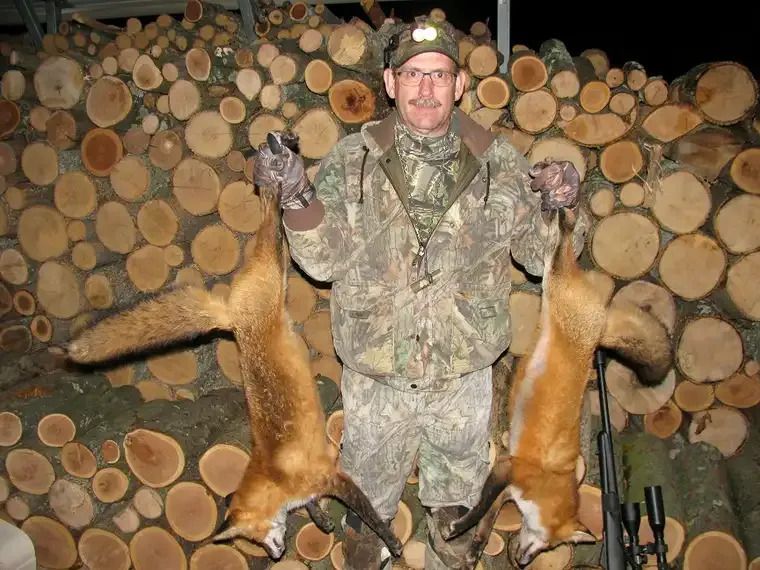 Hunter in camouflage posing with two deer legs in front of stacked cut logs.