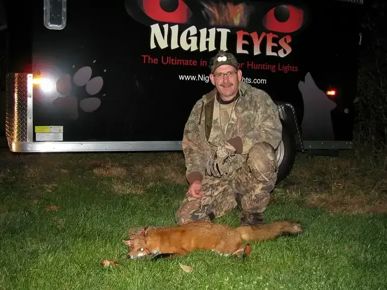 Hunter in camouflage crouches beside a fox on grass in front of a night hunting sign.