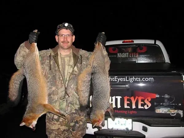 Man in camo holding two large raccoons beside a truck with Night Eyes Lights branding