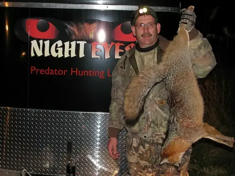 Man posing with two large hunted animals beside a NIGHT EYE predator hunting trailer