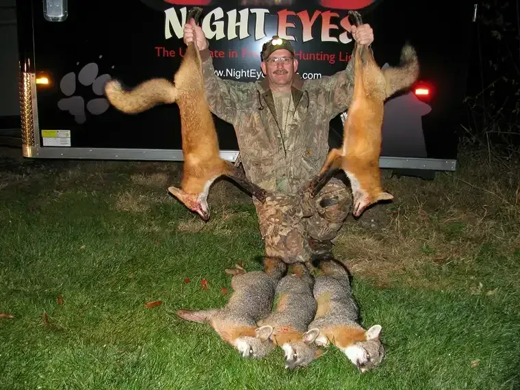 Man holding up two deer and standing by several more on grass at night near a truck.