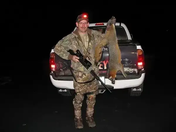Camouflaged hunter holding a rifle and hanging deer beside a pickup truck at night