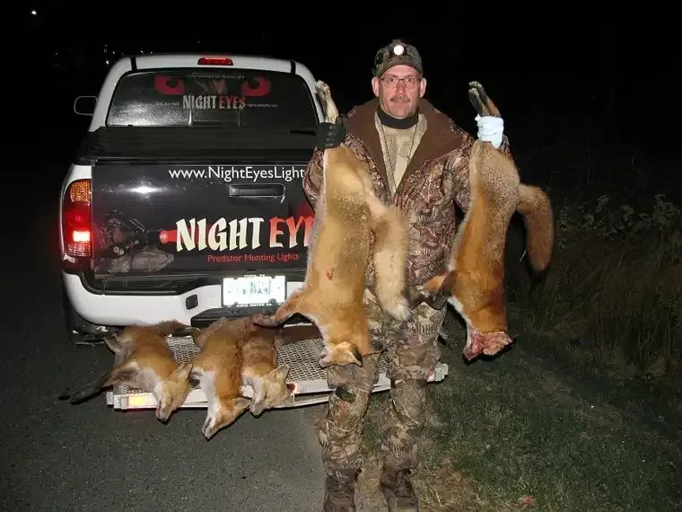 Hunter kneels by a black truck at night, displaying two fox pelts and a fox carcass on the ground.