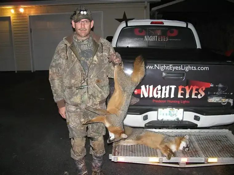 Camouflaged hunter posing with two foxes beside a truck at night