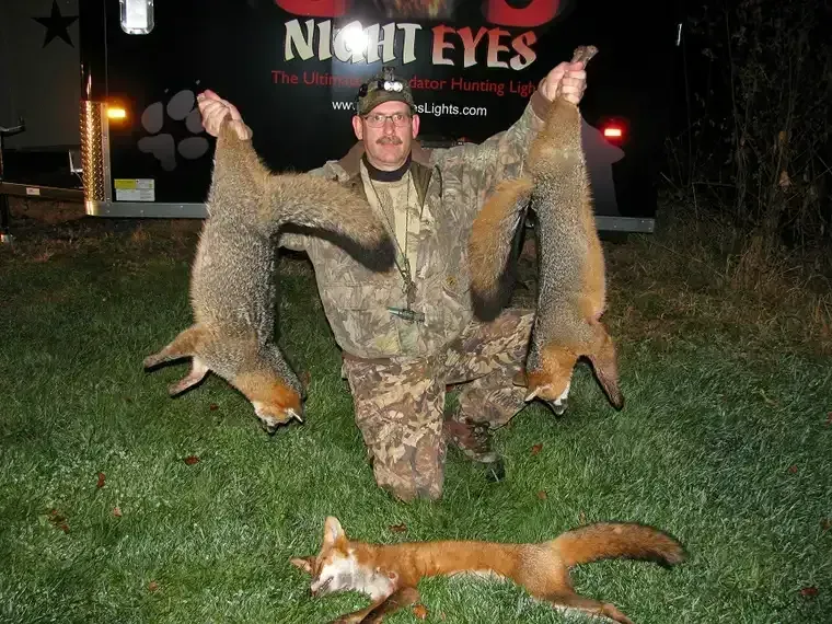 Hunter kneeling at night holding two coyotes, with a fox lying on the grass behind him