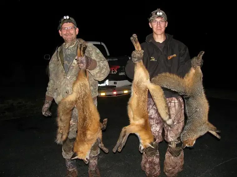Two hunters in camo holding fur pelts and rifles at night beside a vehicle
