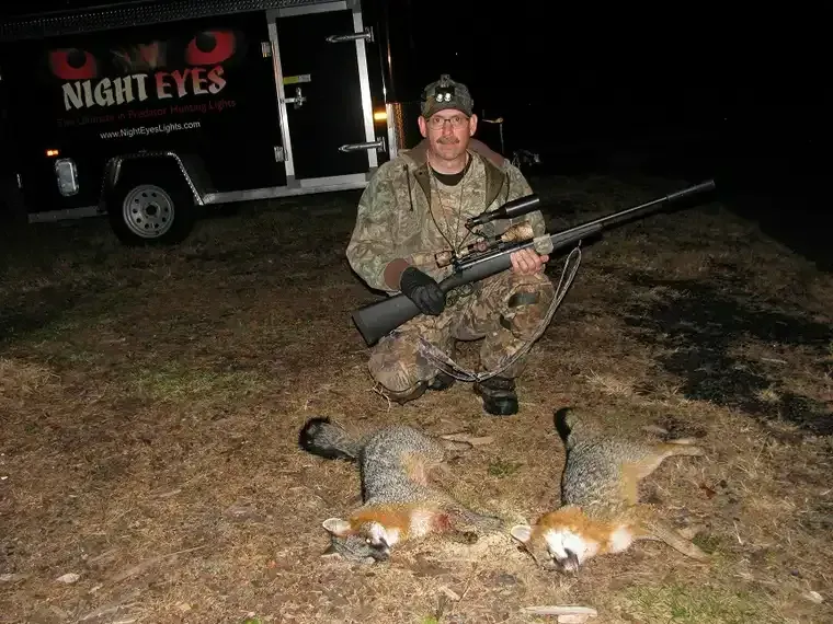 Hunter crouches at night beside two dead foxes, holding a rifle near a trailer marked “NIGHT EYES.”