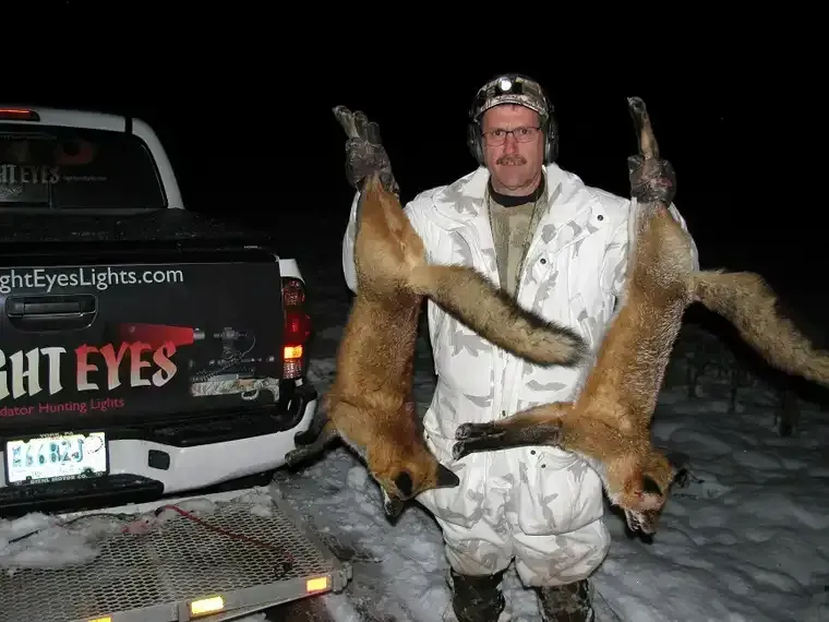Man in white winter camouflage holding two dead foxes beside a truck at night