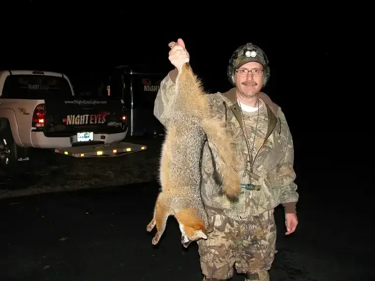 Hunter in camouflage holding a large fox by the tail at night beside a pickup truck