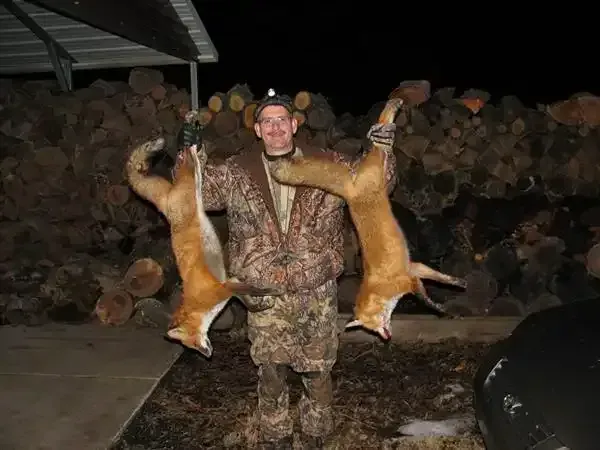 Hunter holding two dead foxes in camouflage, standing in a dark outdoor shed.