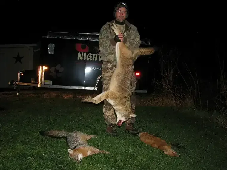 Hunter holding a large coyote beside a trailer, with several other coyotes on the ground at night