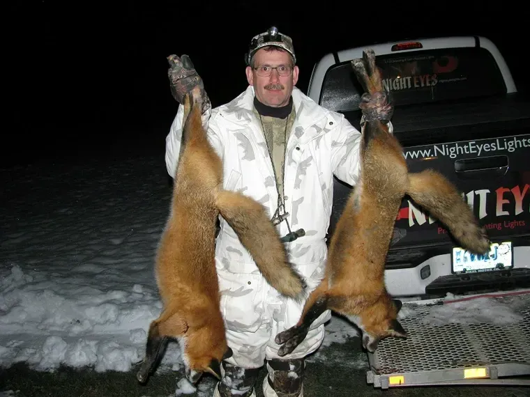 Person in white winter gear holding two large foxes beside a truck at night in the snow