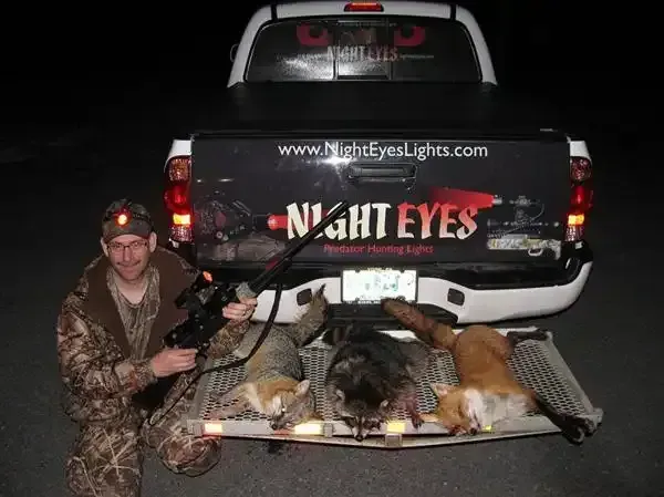 Hunter kneels beside two dead deer and a rifle, posing behind a pickup truck at night