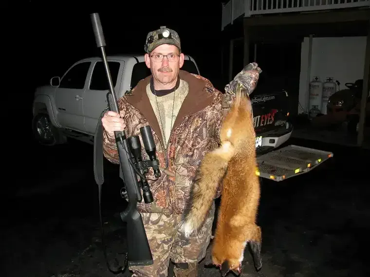 Hunter holding a rifle and a dead fox beside a pickup truck at night