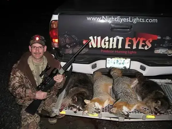 Hunter kneels beside a truck bed full of coyotes, holding a rifle under a Night Eyes logo.
