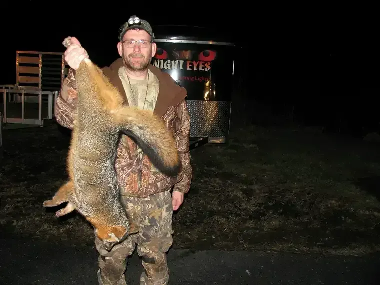 Hunter posing with a large fox he’s holding by the tail at night, wearing camouflage outdoors.
