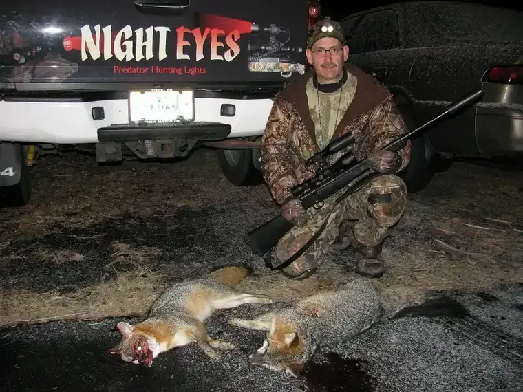 Hunter kneels by a truck beside two foxes, holding a rifle at night.