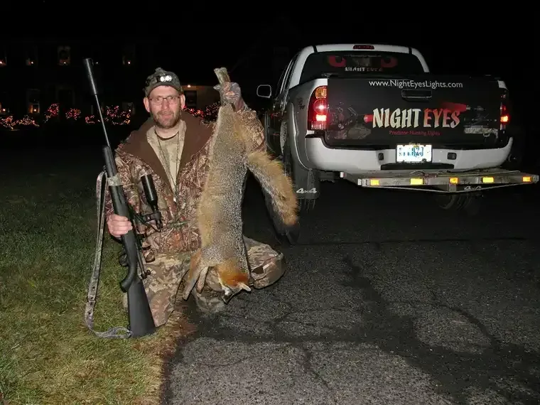Hunter kneels beside a dead coyote, holding a rifle, with a truck labeled “NIGHT EYES” behind him.