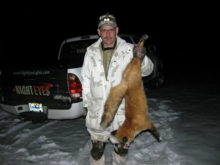 Man holding a hanging fox beside a black truck in the snow at night