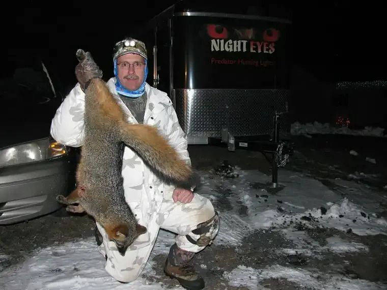 Person in white camouflage kneels beside a fox, holding it up in a snowy night scene.
