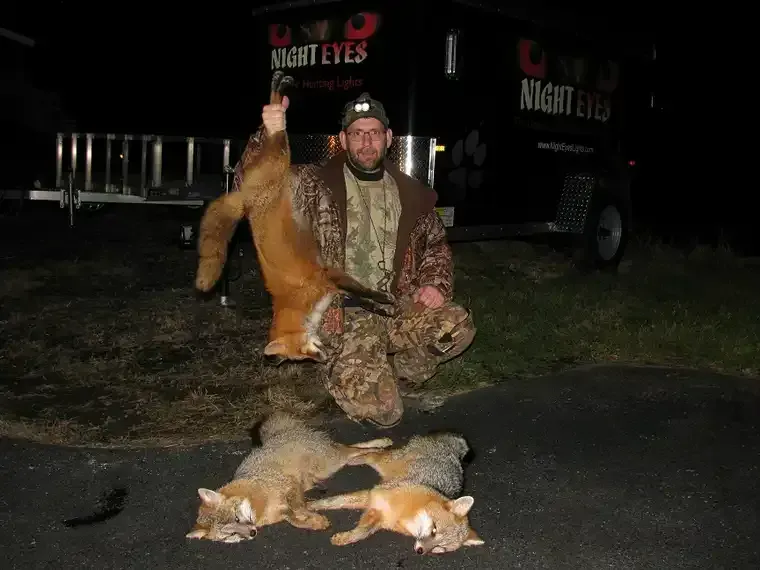Hunter kneeling with several dead foxes on the ground at night beside a truck