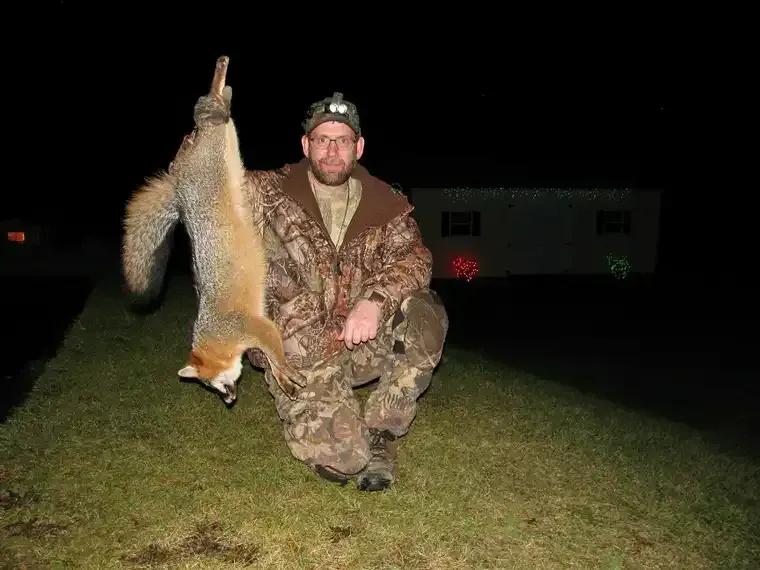 Man kneeling at night, holding up a fox by the tail in a grassy field, wearing camouflage.