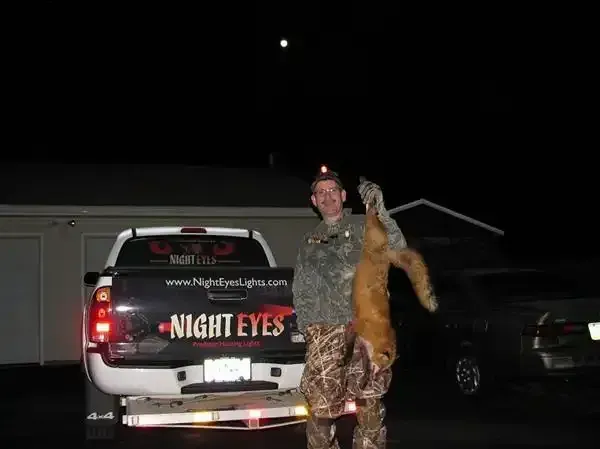 Person in camouflage holds a deer beside a pickup truck at night, with headlights and garage in the background