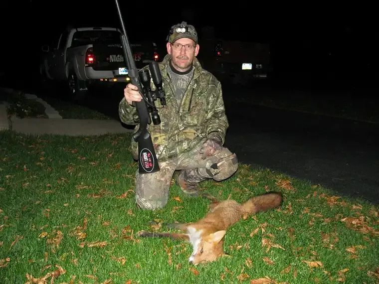 Hunter kneeling on grass at night beside a rifle and a dead fox, with trucks in the background