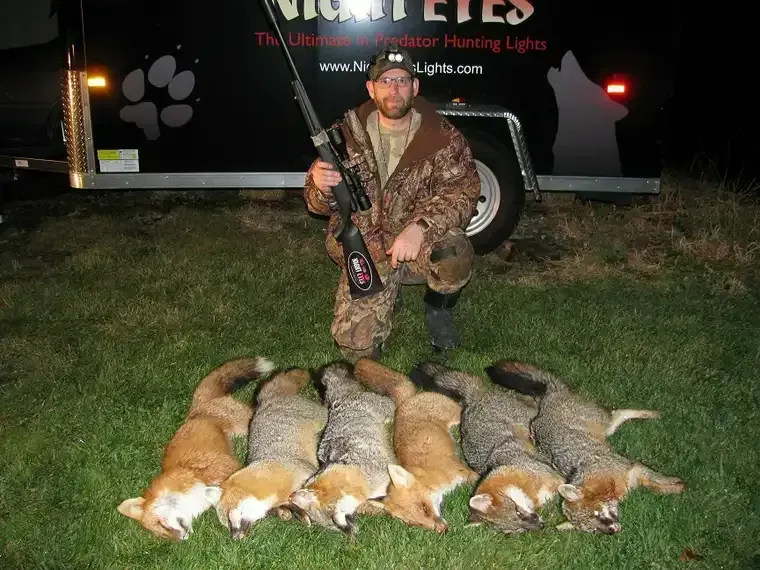 Hunter kneeling behind a row of dead ducks beside a hunting trailer at night
