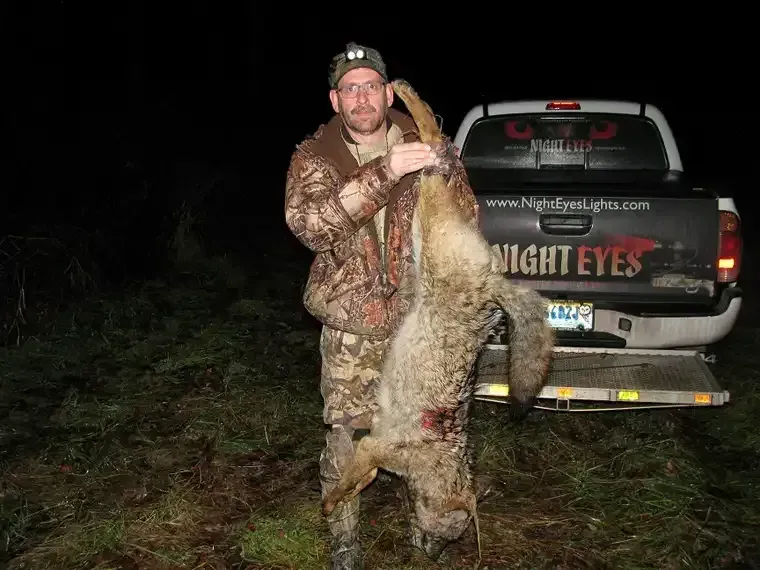 Hunter holding a large alligator at night beside a pickup truck in a muddy field