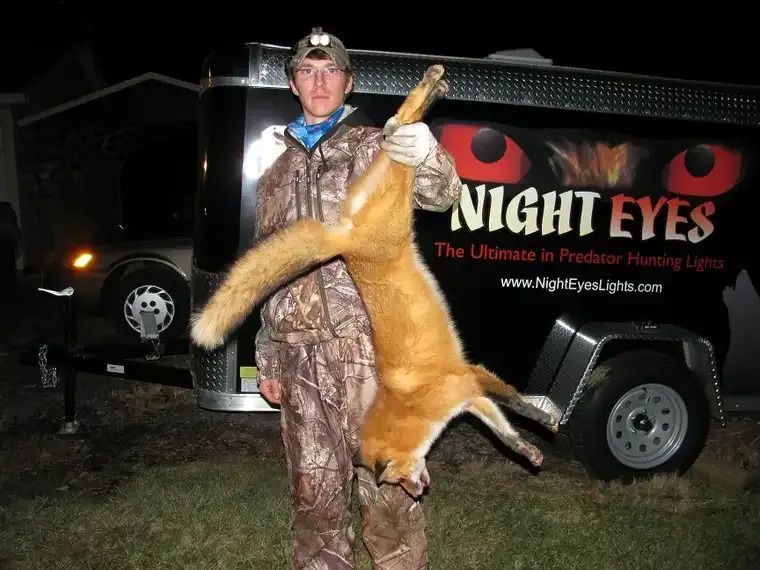 Hunter holding a large fox beside a “Night Eyes” trailer at night