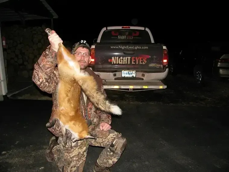Man in camouflage holding up a large fox at night in a driveway, with a pickup truck behind him.