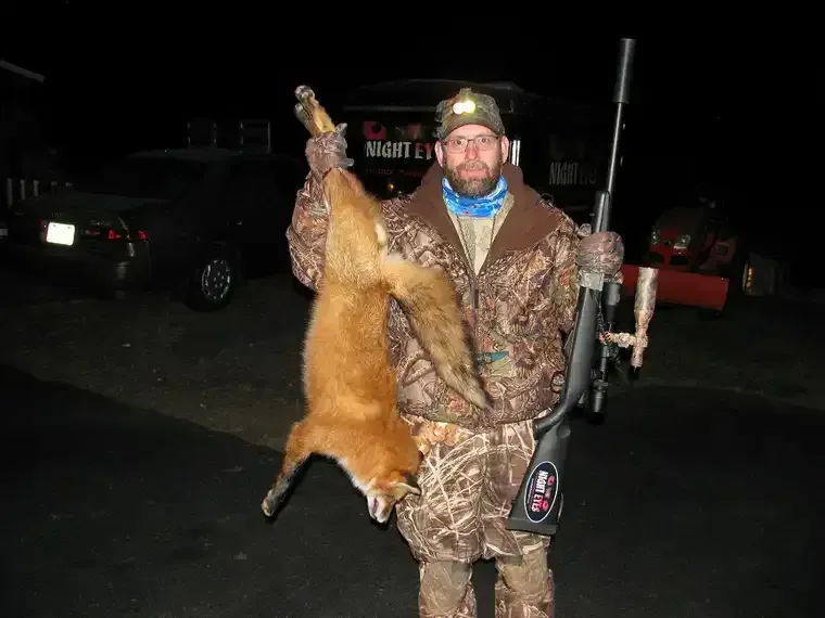 Hunter in camouflage holding a hanging fox and a rifle at night outdoors