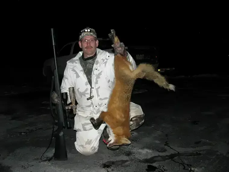Person in white camouflage kneels beside a deer at night, holding a rifle in a snowy field.