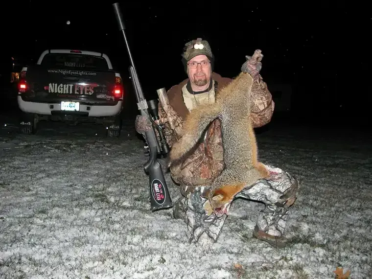 Hunter kneeling at night, holding a large fur pelt and rifle beside an SUV on snowy ground
