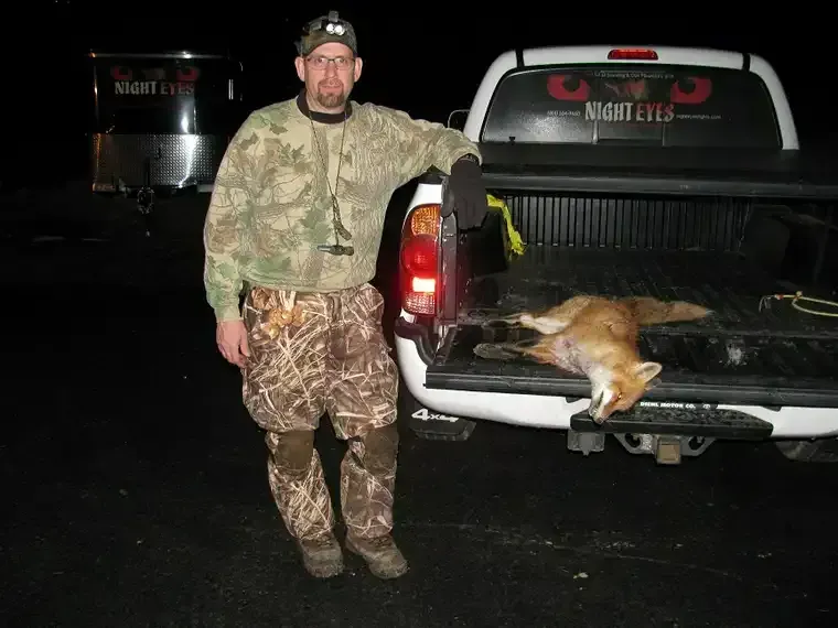 Man in camouflage stands by pickup truck with a fox or coyote in the open tailgate at night