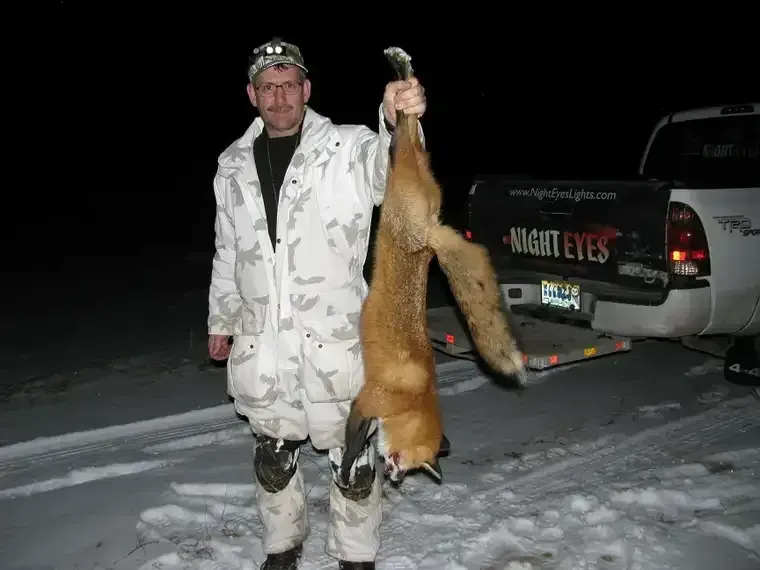 Man in white camo holding a large fox beside a truck in the snow at night
