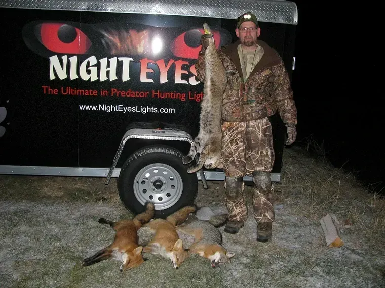 Man standing beside a Night Eyes trailer with two dead foxes on the ground at night