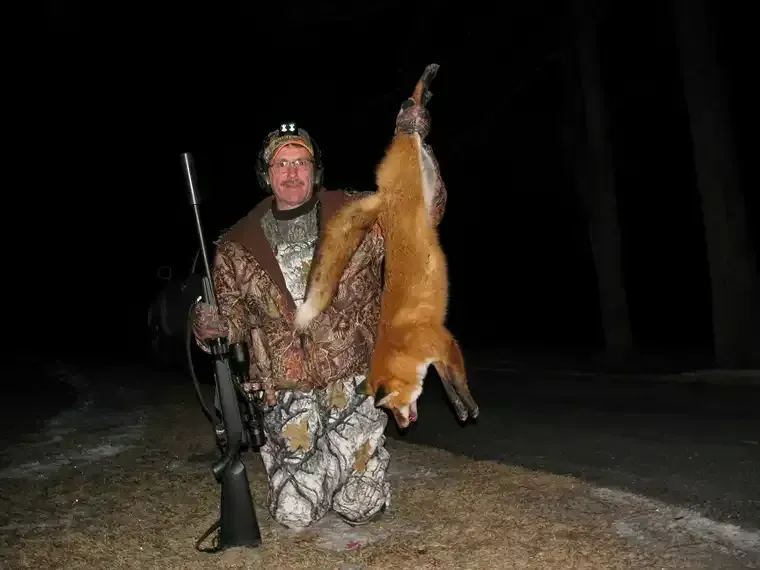 Hunter in camouflage holding a rifle beside a hanging fox at night