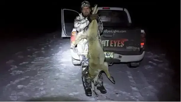 Person holding a large fish beside an SUV at night on snowy ground, with a spotlight illuminating the scene