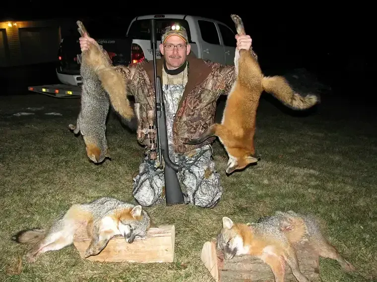 Man kneeling at night with four dead foxes, holding two by the tails, beside a truck on grass.